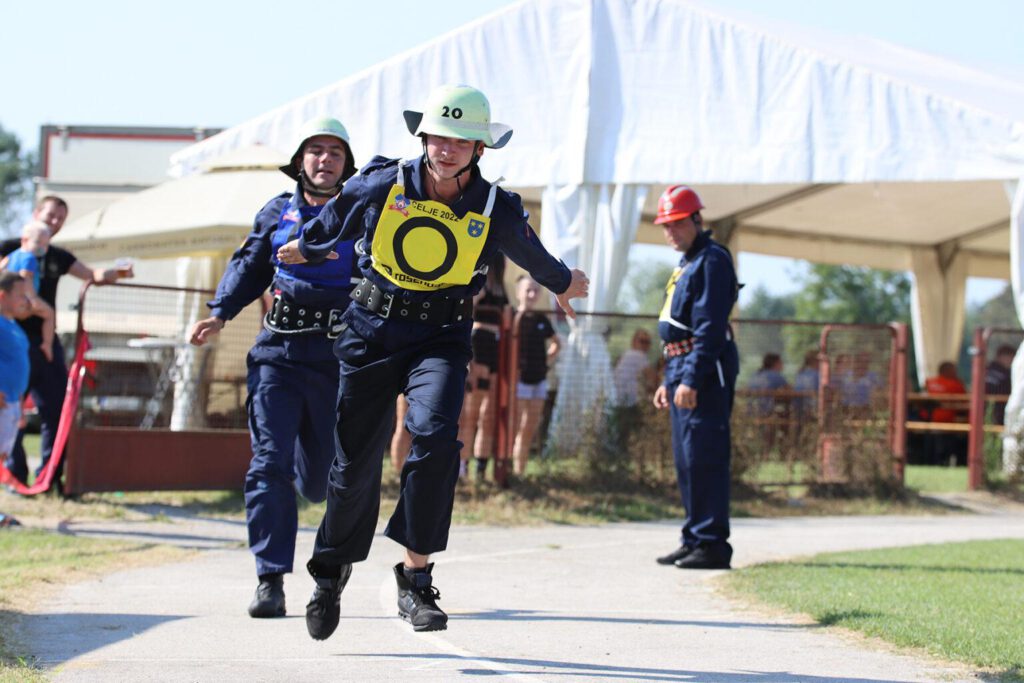 Im Vordergrund läuft eine Person in blauer Uniform und Helm auf einer Rundbahn. Sie läuft los, während eine zweite Person ihr einen Staffelstab übergibt.