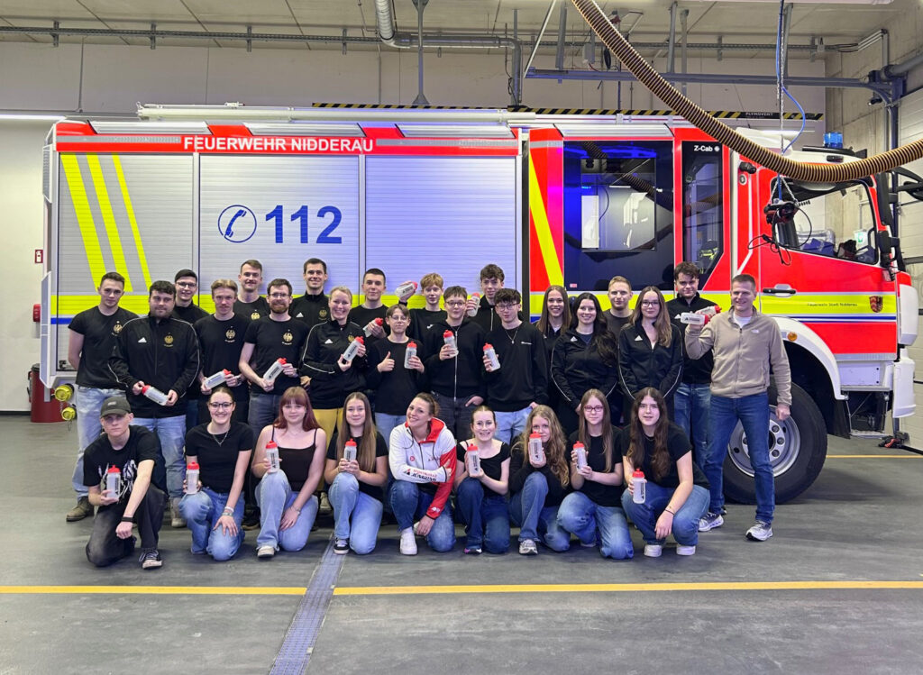 Eine Menschengruppe mit schwarzen Trainingsjacken steht in einer Gerätehalle vor einem Feuerwehrlöschfahrzeug. Die Personen halten Trinkflaschen in die Kamera.