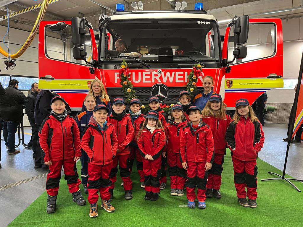 Ein Dutzend Kinder mit roten Feuerwehranzügen steht vor einem Feuerwehrauto, auf dessen Motorhaube eine Blumengirlande befestigt ist. Das Foto wurde bei einer Feierlichkeit aufgenommen