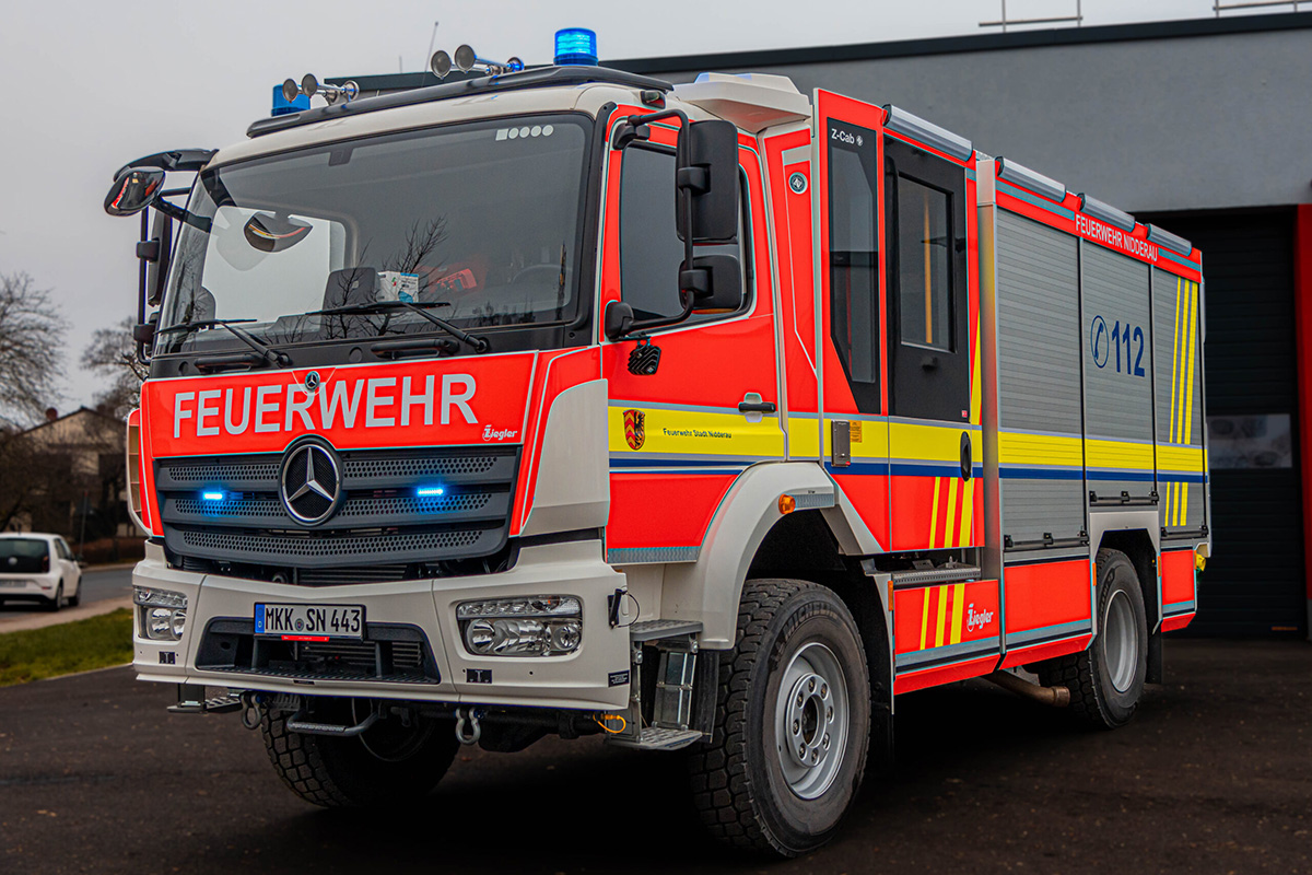 Ein modernes, rot lackiertes Löschgruppenfahrzeug der Feuerwehr steht auf einem asphaltierten Platz vor einem Gebäude mit grauem Tor. Das Fahrzeug trägt auffällige gelbe und weiße Reflexstreifen sowie die Aufschrift 'FEUERWEHR' in großen weißen Buchstaben auf der Front.
