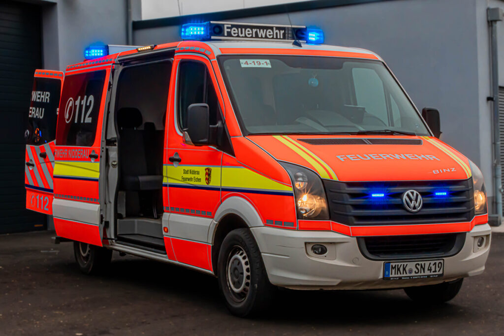 Ein orange-rotes Einsatzfahrzeug mit gelben und weißen Streifen steht auf einem asphaltierten Untergrund. Auf dem Dach sind mehrere blaue Rundumleuchten montiert. Die Schiebetür auf der rechten Seite ist geöffnet. Auf der Motorhaube und an der Seite steht in weißen Großbuchstaben das Wort 'Feuerwehr'.