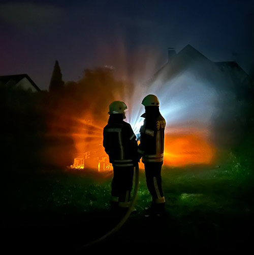 Zwei Silhouetten von zwei Feuerwehrleuten in der Nacht. Sie richten einen Schlauch auf ein orange-blau beleuchtetes Objekt.